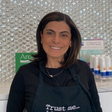 Ingrid, passionate owner, in her salon uniform in front of the Aroma Waxing Clinic logo, radiating expertise and a commitment to client confidence